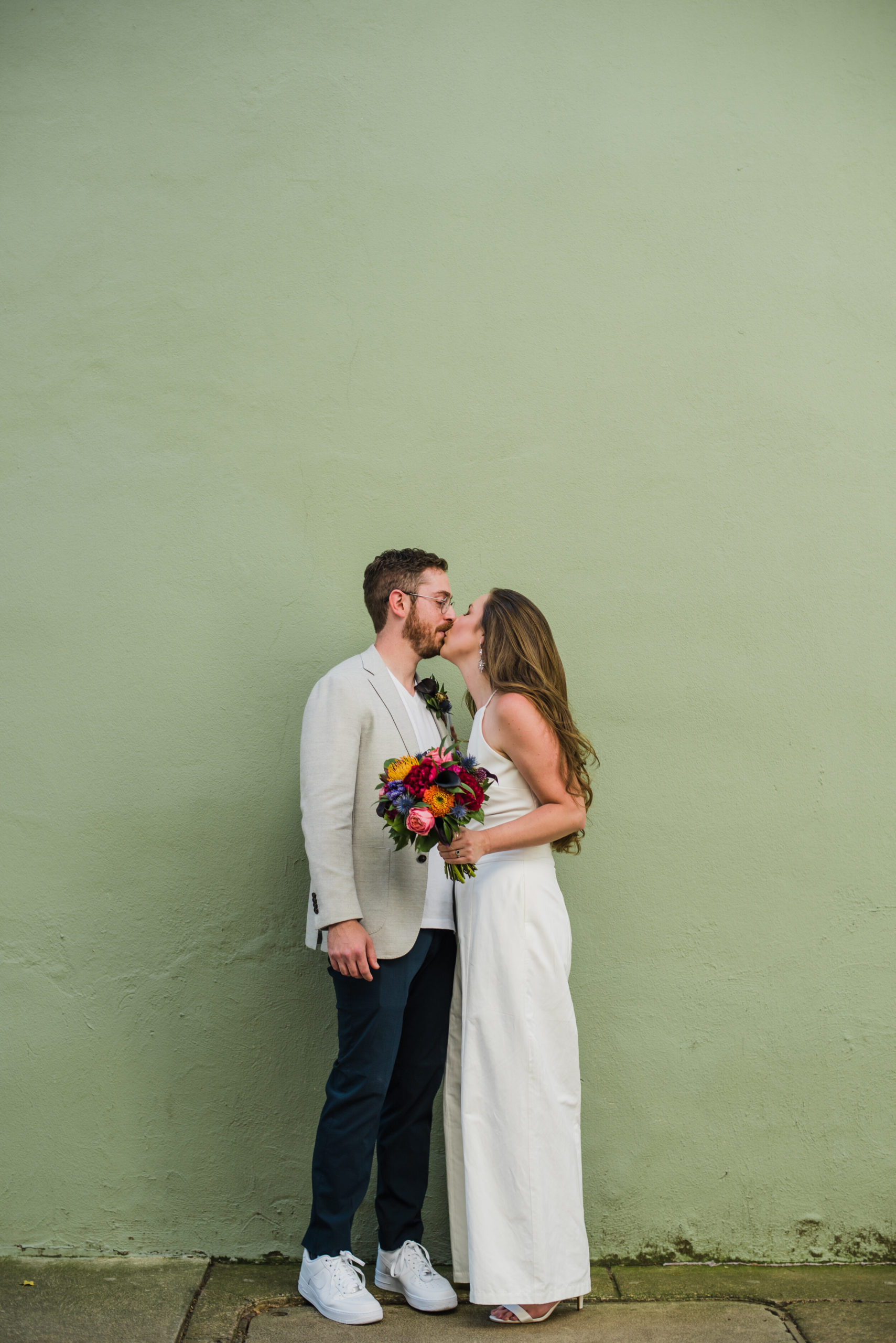 Wedding couple kissing in front of a pale green wall.