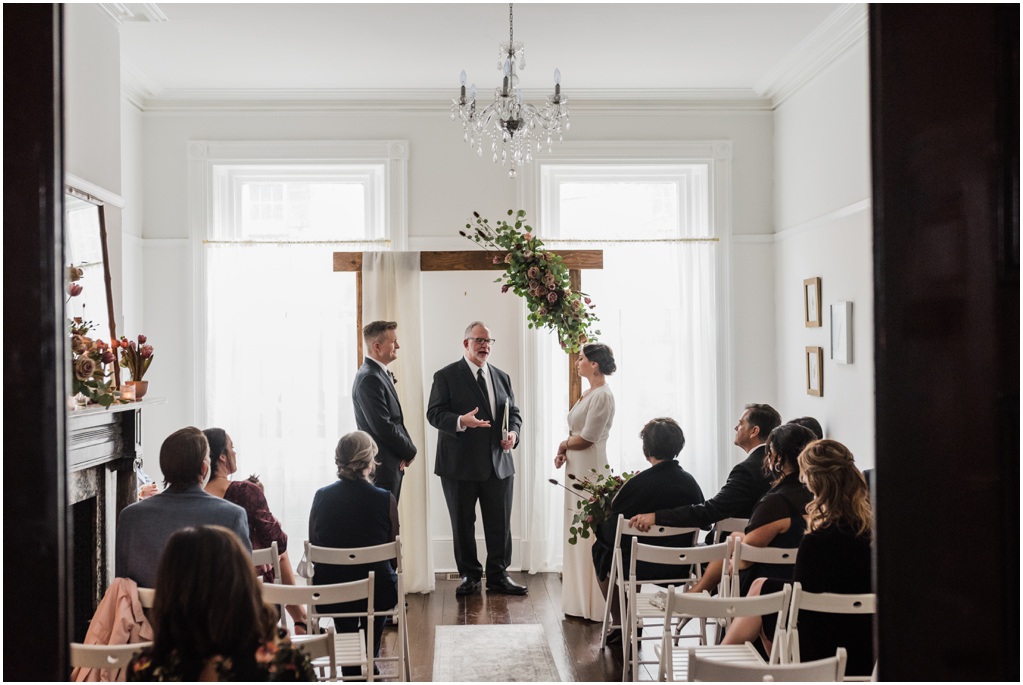 A wedding ceremony under an arbor with florals and white fabric inside a house