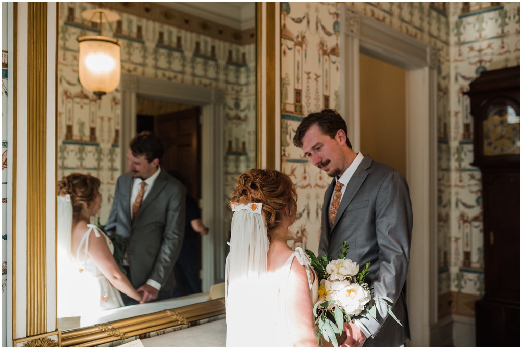 An interior photo with the couple standing by a mirror in front of patterned wallpaper
