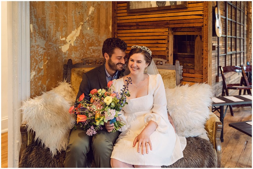 A couple sits in their wedding venue on a couch with white fur pillows.