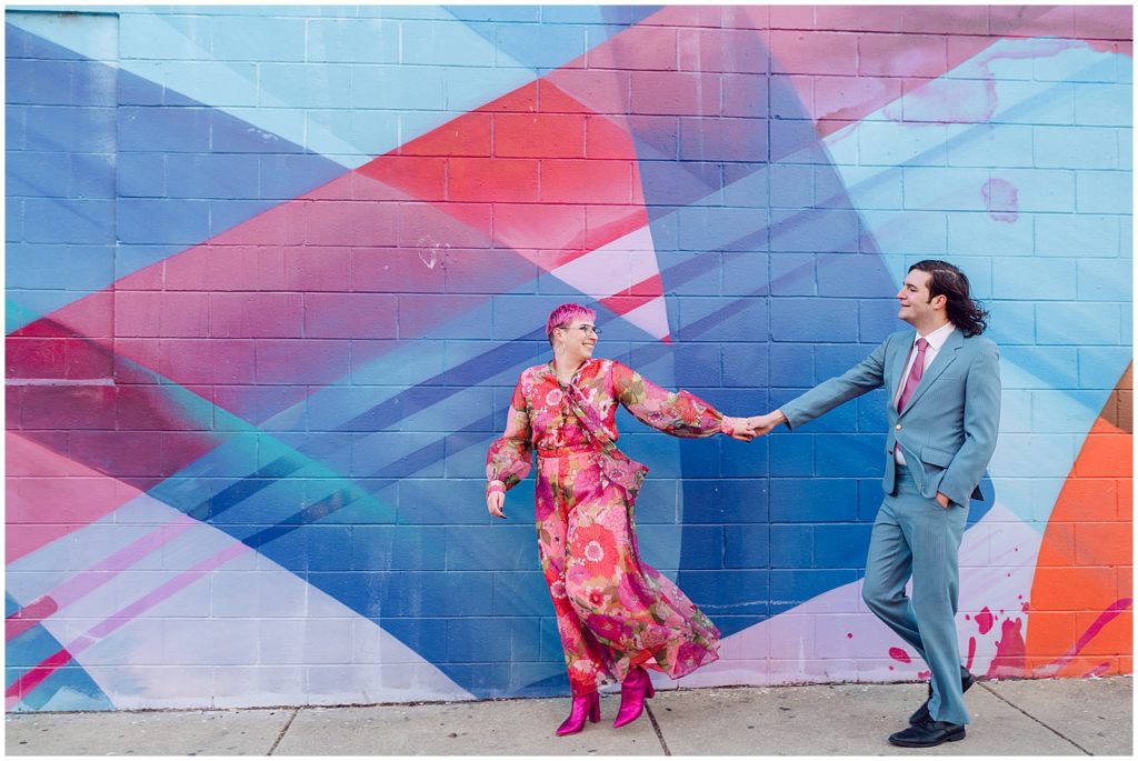 A couple in colorful wedding attire walks past a blue and pink mural.