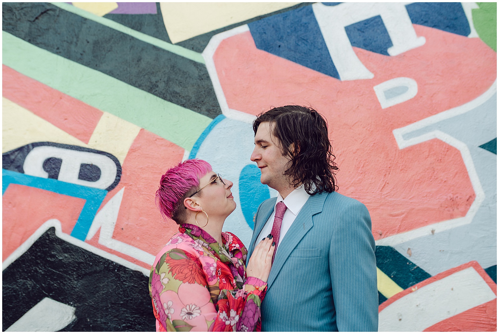 A couple in colorful wedding attire stands in front of an abstract mural.