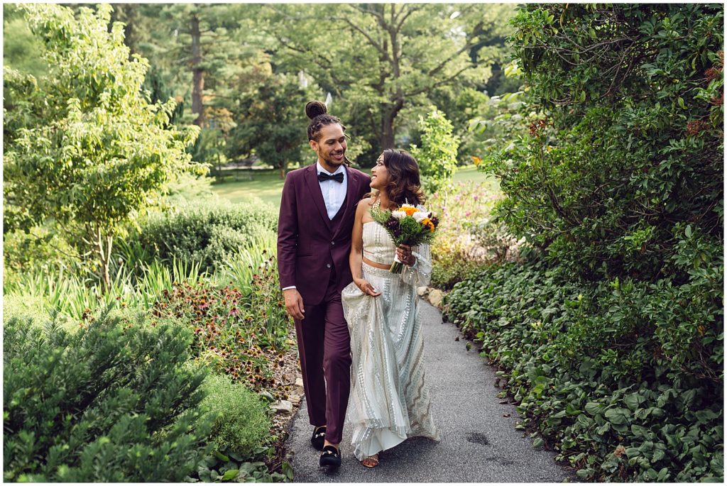 A couple wearing white and burgundy walks down a wooded path at their Tyler Arboretum wedding.