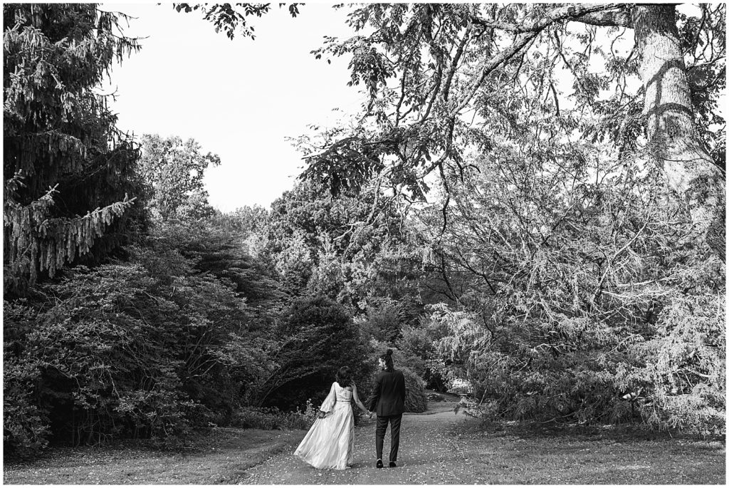 A couple walks under the trees before their Tyler Arboretum wedding.