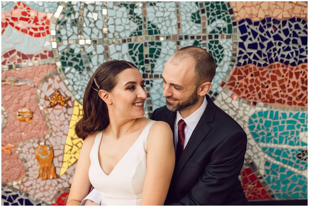 A couple smiles in front of a mosaic wall.