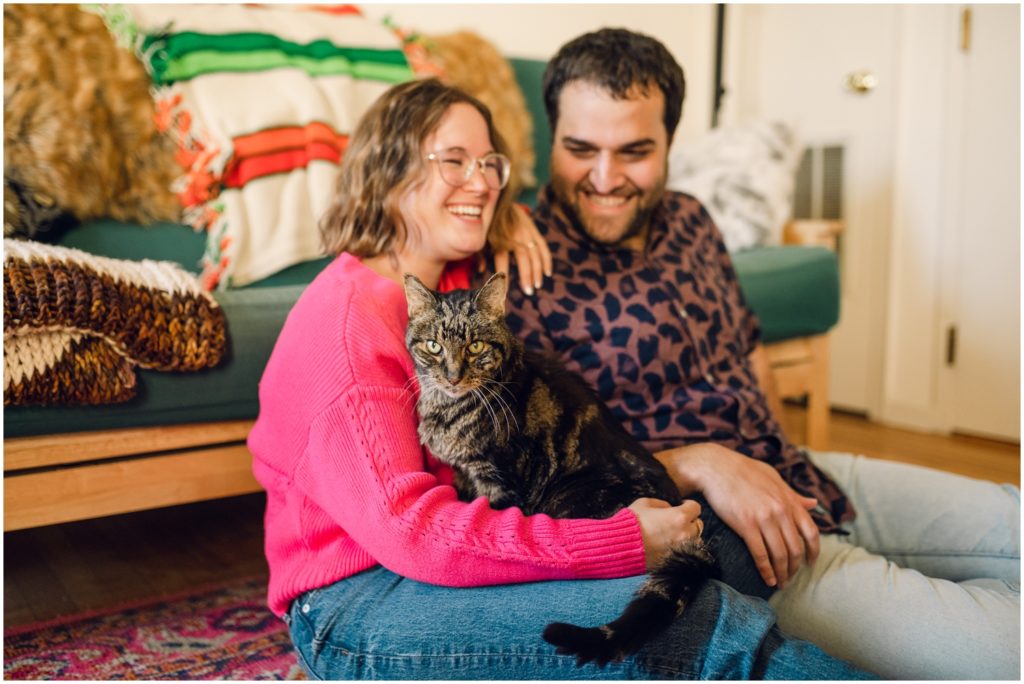 A couple holds their cat and smiles during Philadelphia engagement photos in their home.