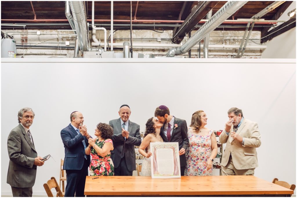 Eliana and Marcus kiss and hold up their ketubah while family members clap.