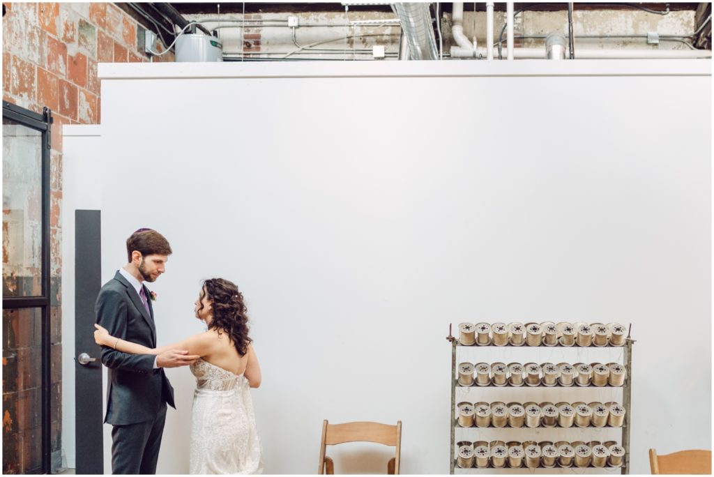 Eliana and Marcus stand to the side beside a rack of yarn spools at their unique Philadelphia wedding venue.