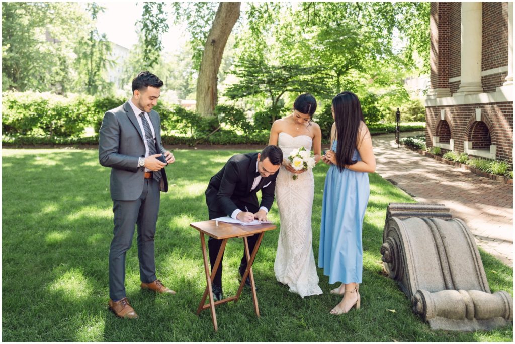 A groom leans over a small table to sign a marriage license in a garden.