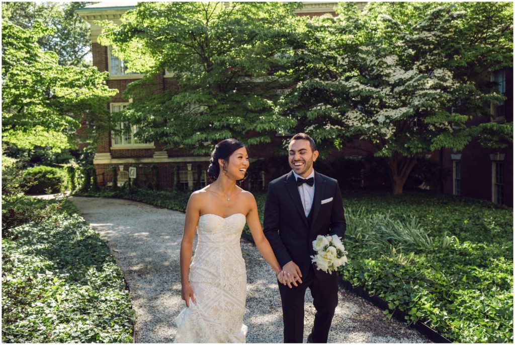 A groom carries a bride's bouquet while they walk through a garden at their alternative to a wedding.