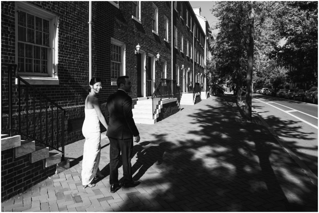A bride and groom walk down a dappled sidewalk holding hands.