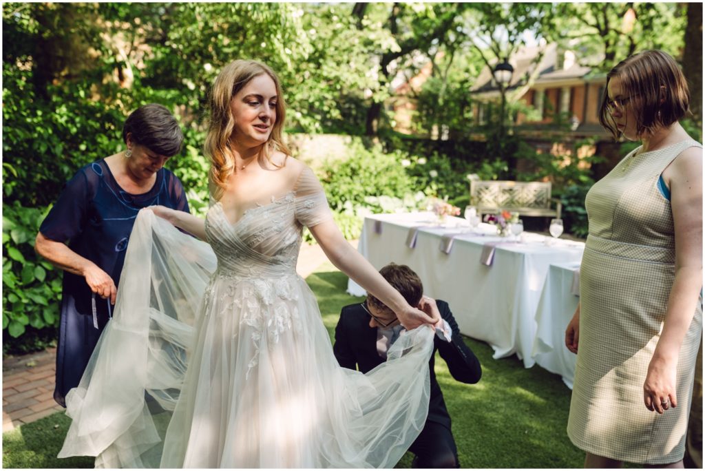 A groom helps bustle a bride's dress at a garden wedding.