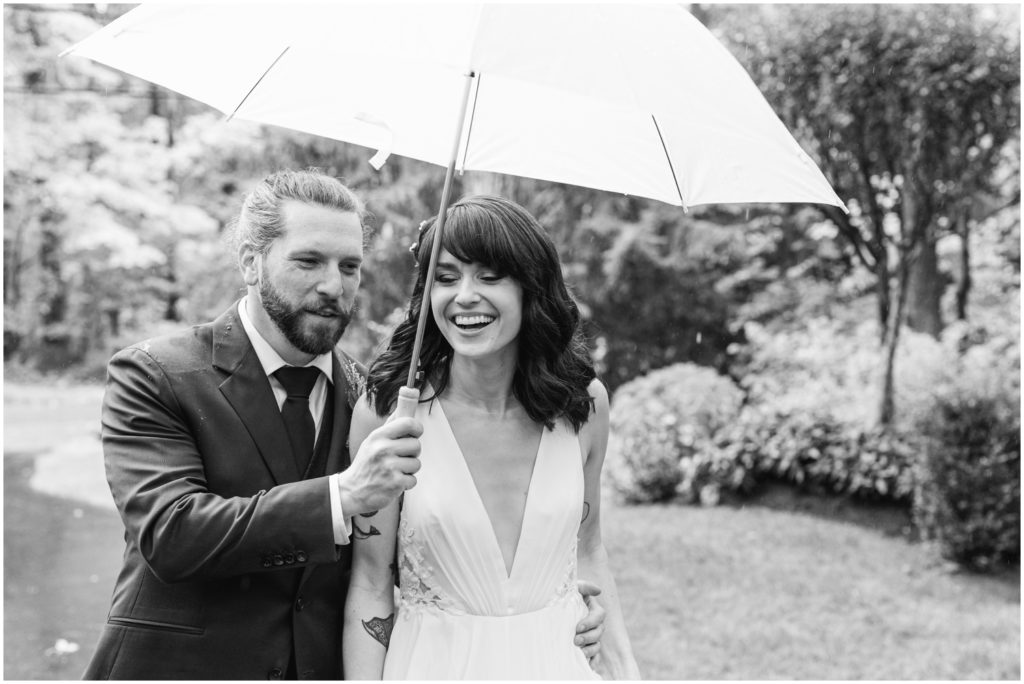 A couple in wedding attire walks across a lawn under a shared umbrella.