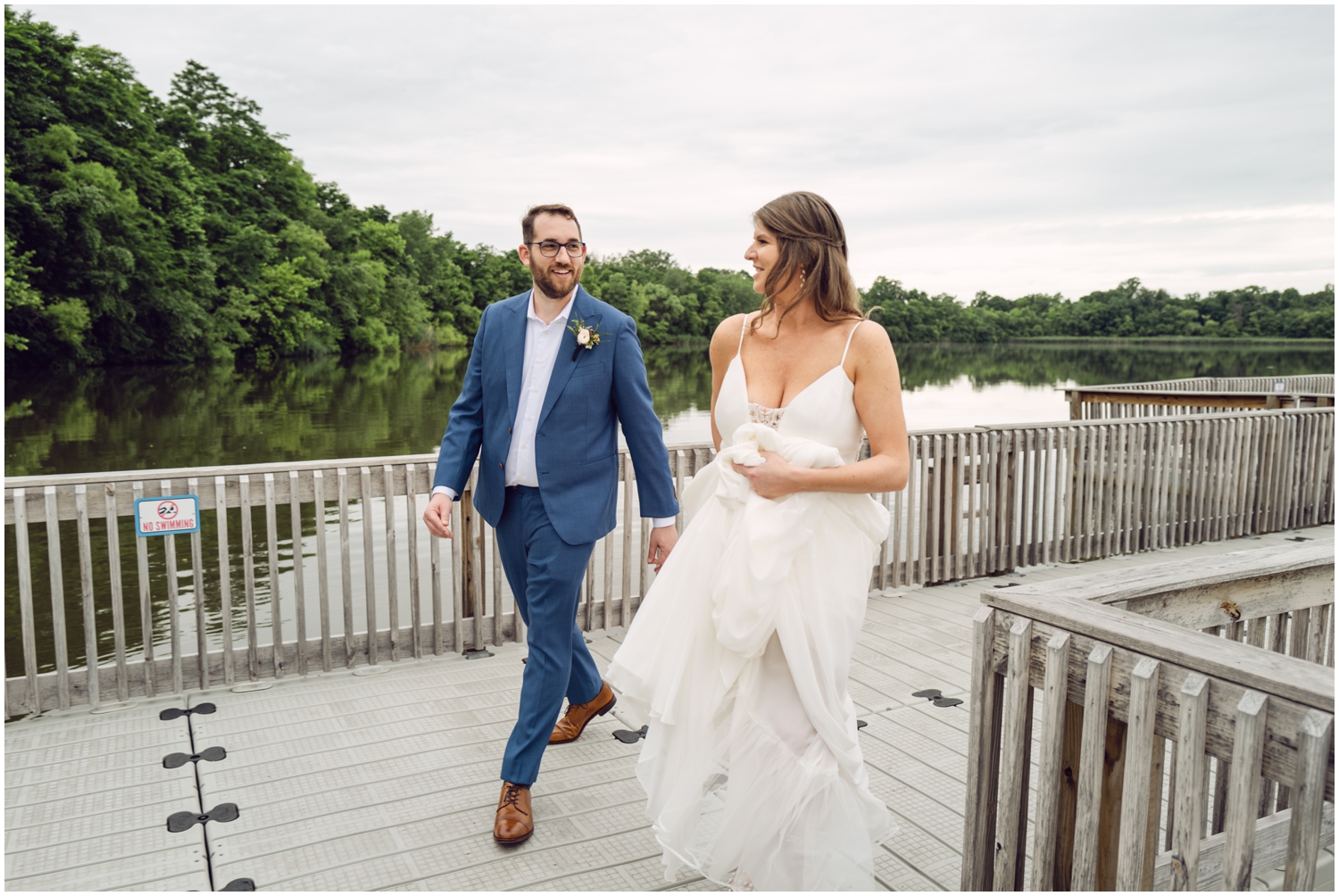 A couple in wedding attire runs up the walkway beside a reservoir.