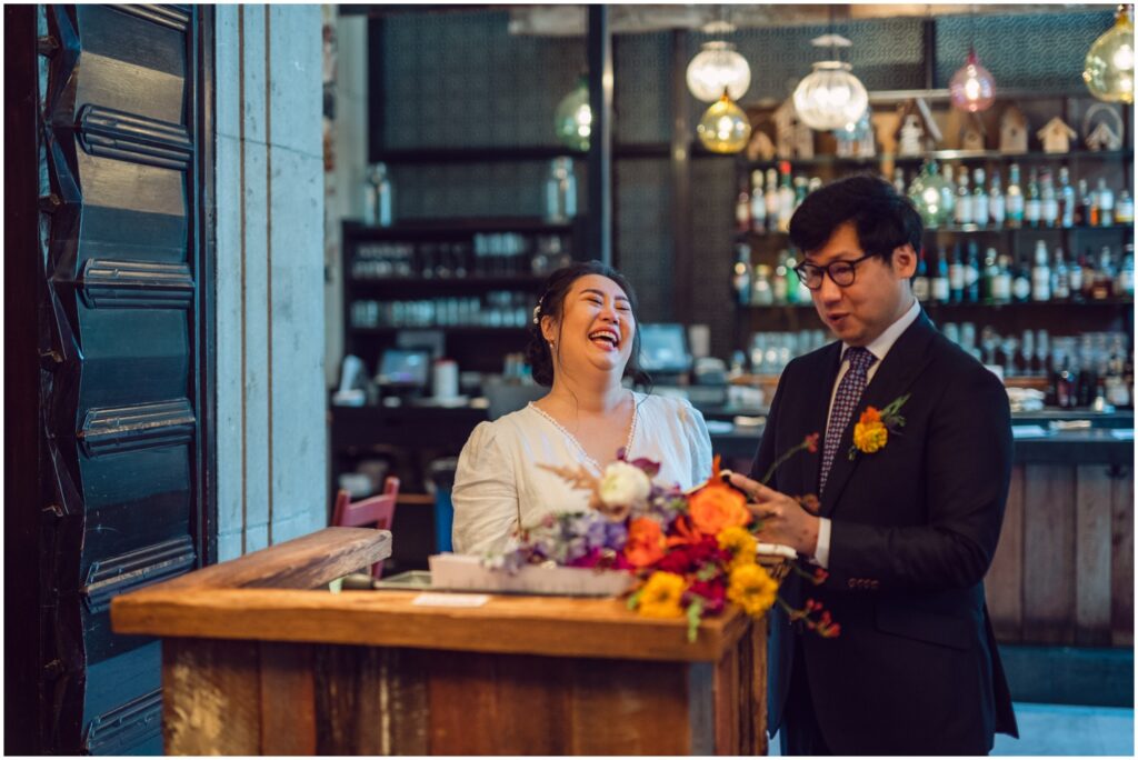 A bride laughs while a groom reads his vows at their restaurant wedding.