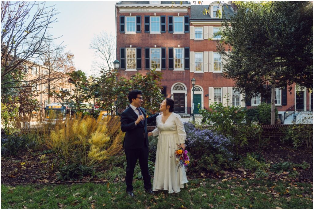 A bride and groom stand in front of flowering bushes in Washington Square.
