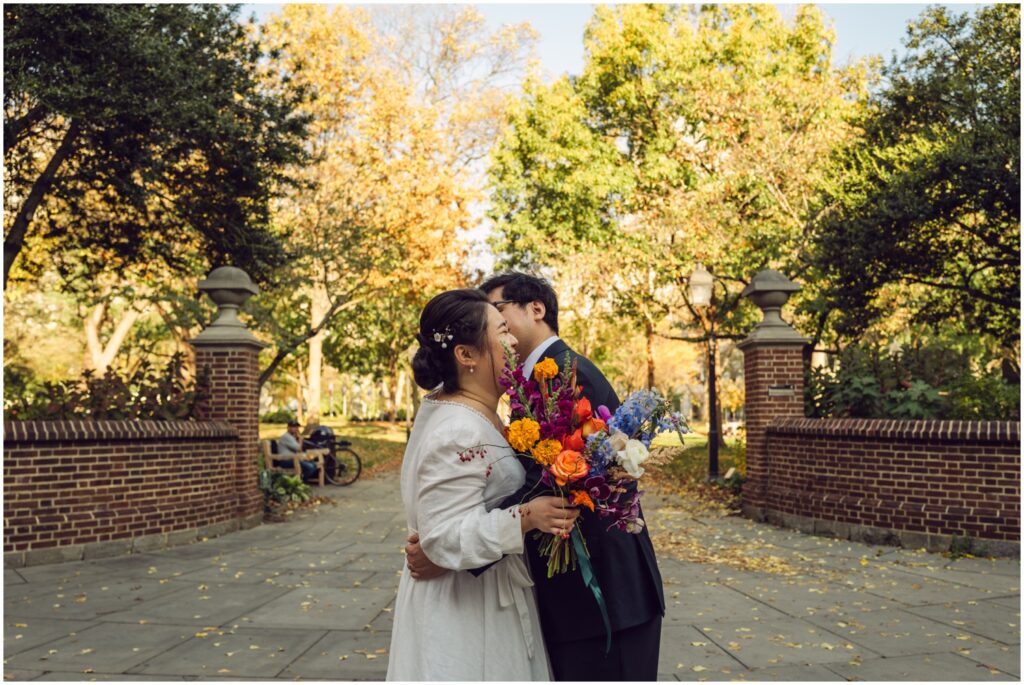 A groom kisses a bride's cheek at the entrance of a park at their Philadelphia wedding.