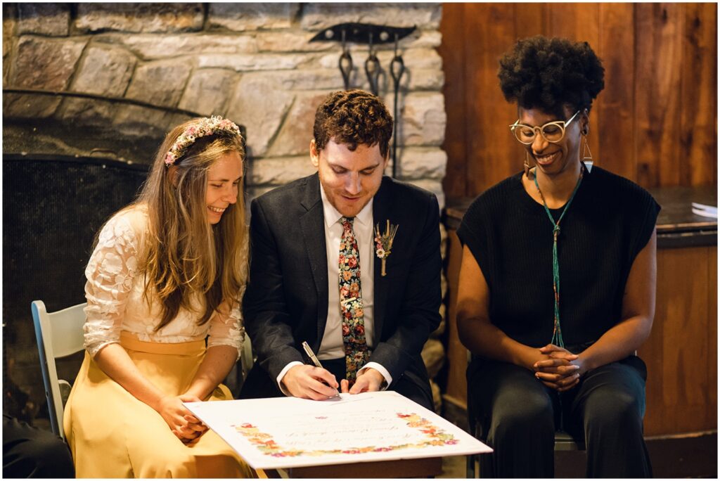 A groom sits beside a bride and signs a Quaker wedding certificate.