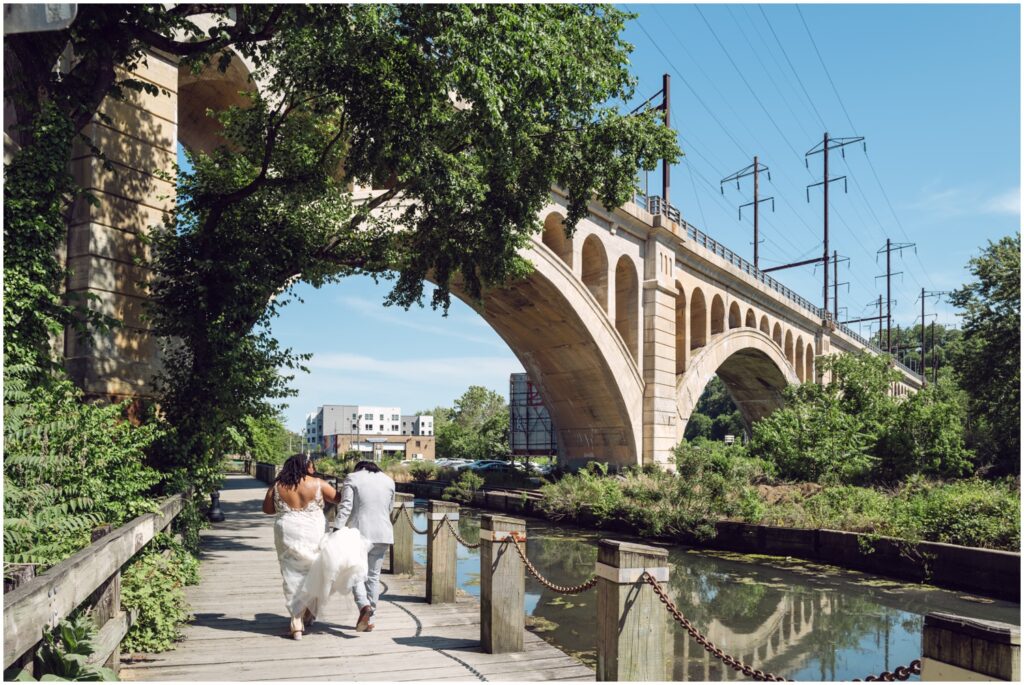 A groom carries a bride's train while they walk through a Philadelphia park beside a river.