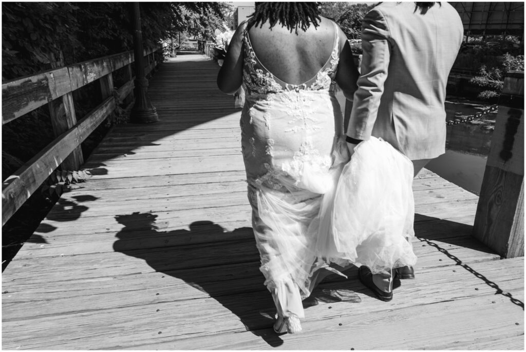 A groom carries a bride's train along a boardwalk.