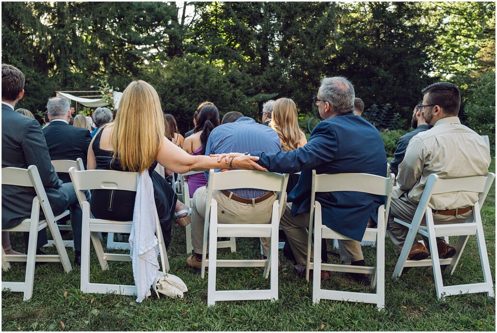 Wedding guests touch each other's hands along the backs of their chairs.
