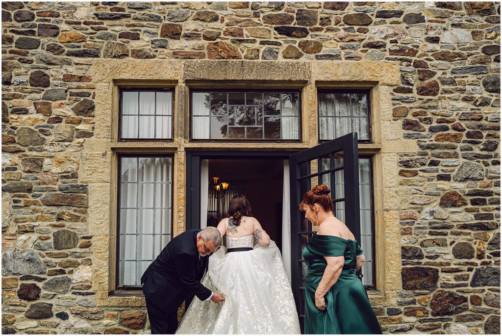 A bride's parents help her carry her bridal gown train through a door.