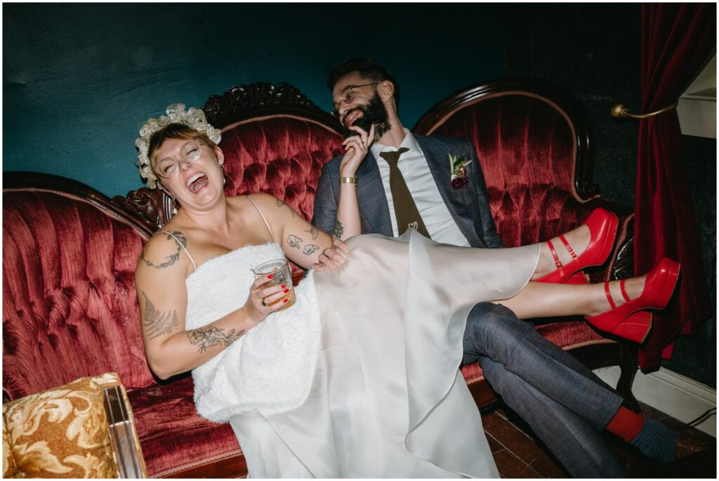 A bride and groom sit on a vintage couch laughing at a Philadelphia wedding venue.