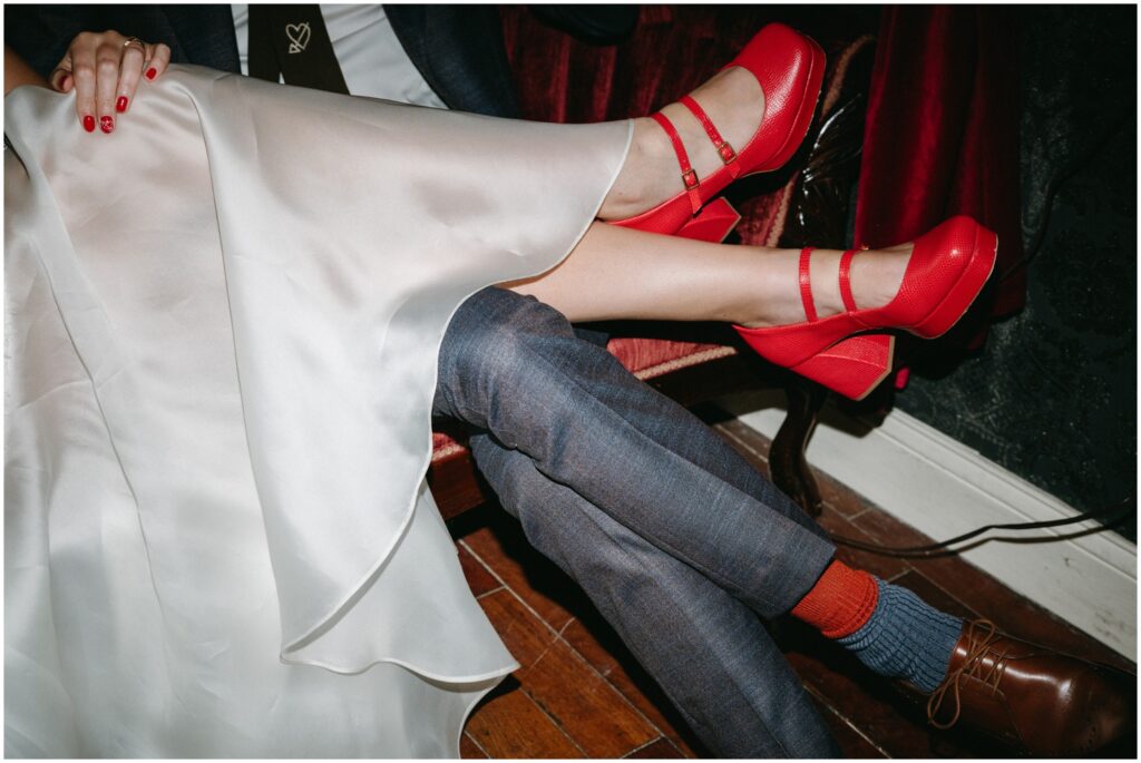 A bride drapes her legs over a groom's and shows off her red wedding shoes.