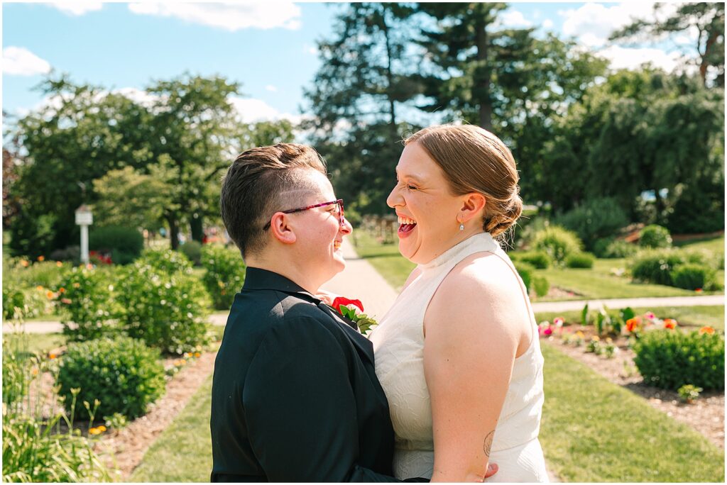 Two women in wedding attire smile at each other in a park.