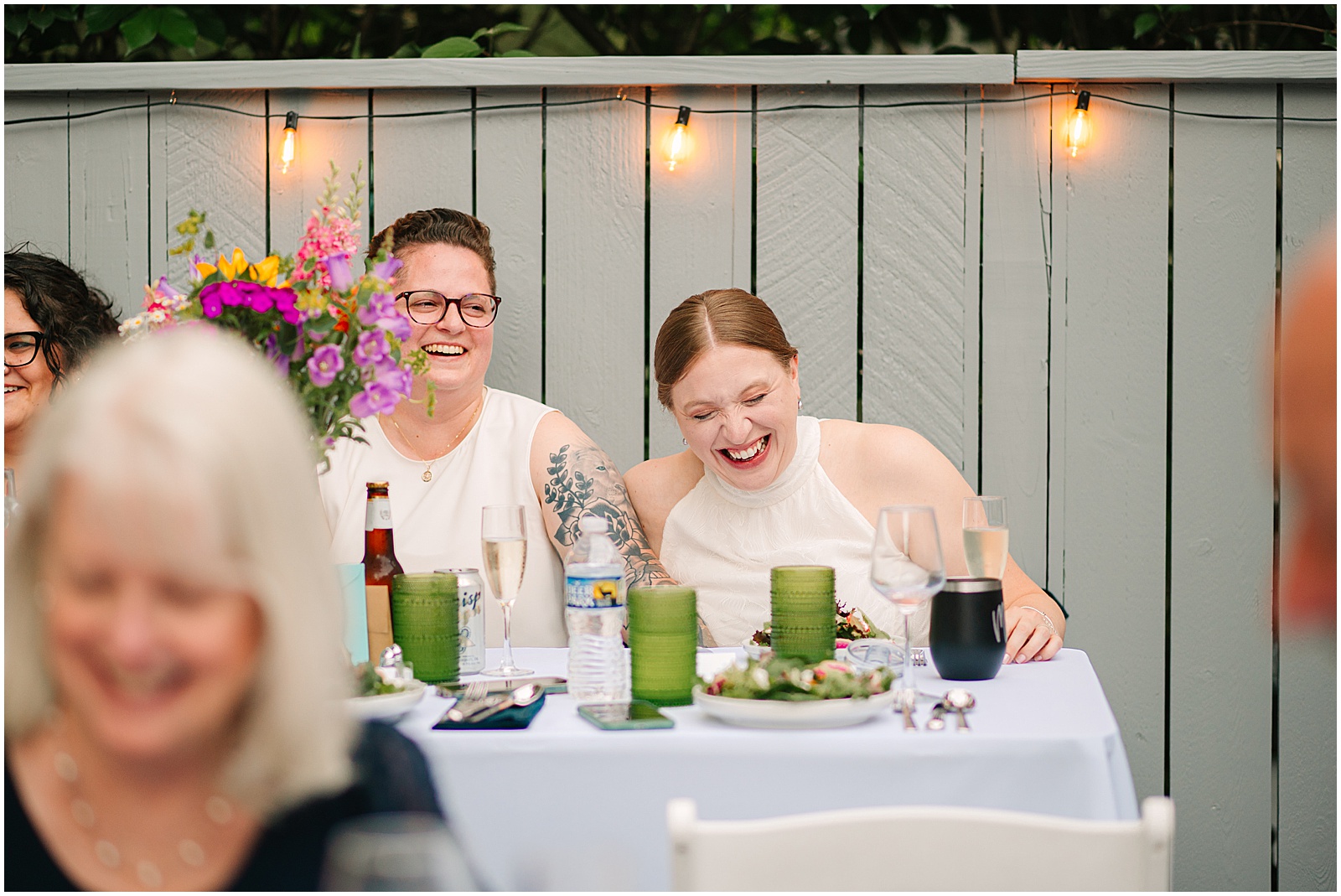 Two brides sit at a table laughing after their Malcolm Gross Rose Garden wedding.