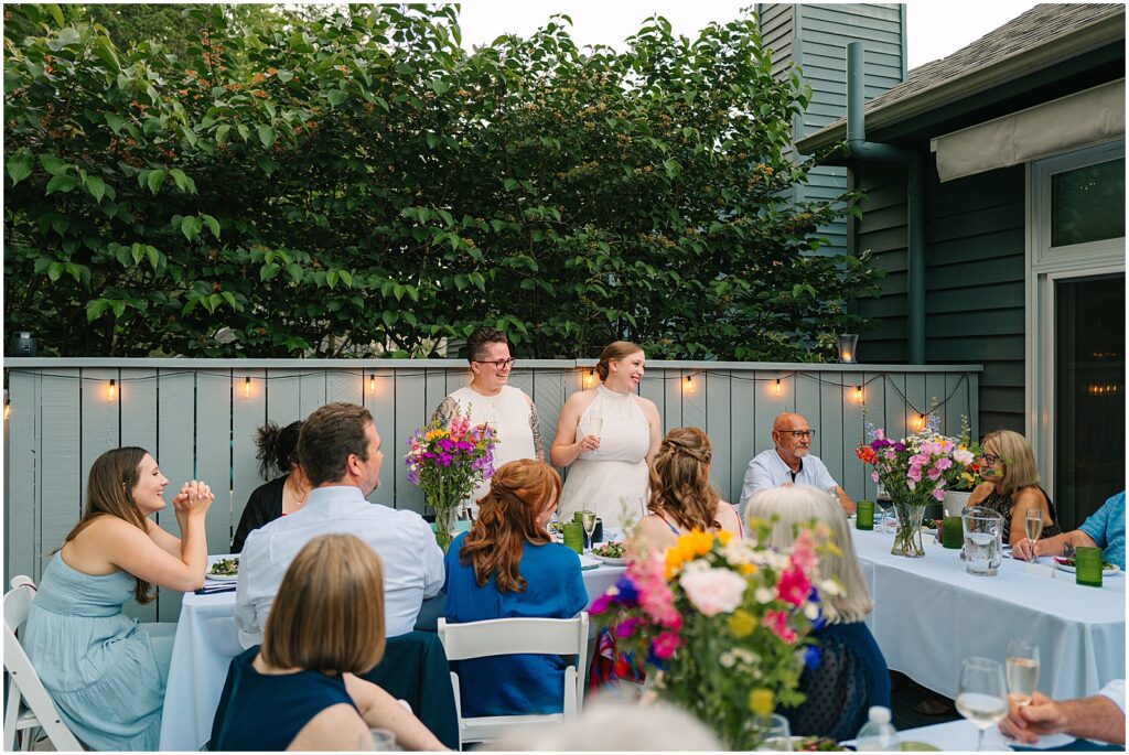 Two brides stand to speak during a backyard wedding reception.