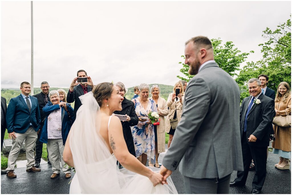 A bride and groom hold hands and smile at each other on a cloudy day.