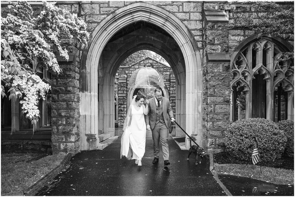 A bride and groom run through the rain under a clear umbrella.