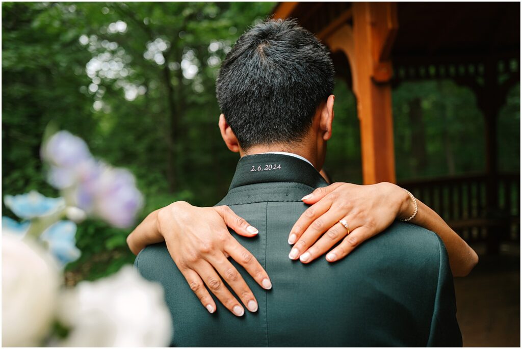 A bride puts her hands around a groom's shoulders, showing her wedding ring.