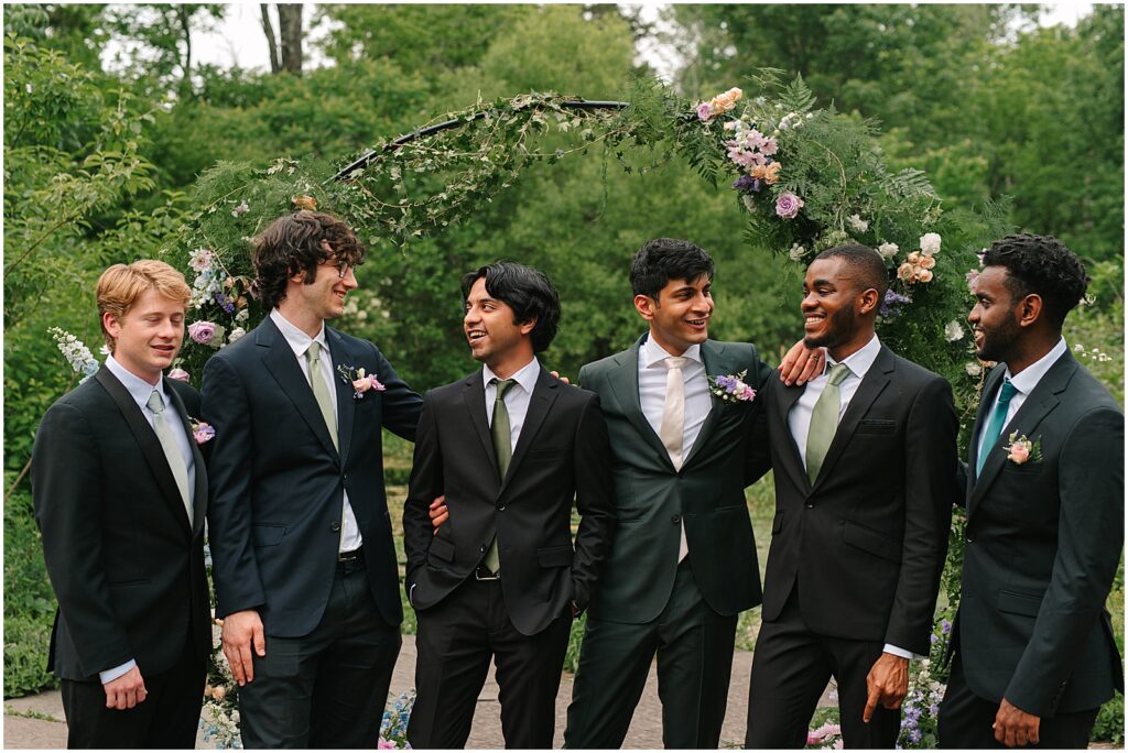 A groom poses with his groomsmen in front of a floral wedding arch.