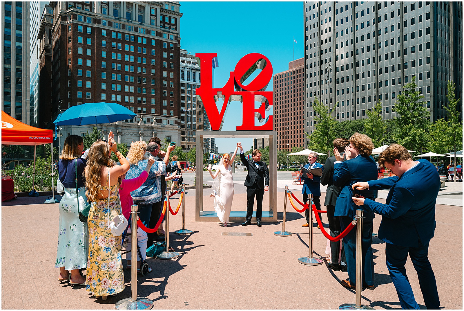 A bride and groom raise their arms as their guests cheer in front of the LOVE sculpture in Love Park Philly.