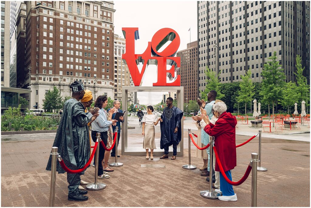 A bride and groom raise their arms as guests cheer in front of the LOVE sculpture at their Love Park Philly wedding.