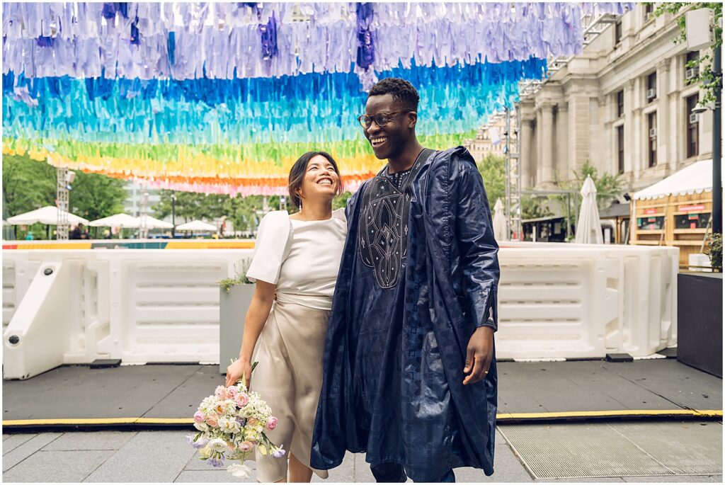 A bride and groom smile as they stand beneath an installation of colorful ribbons in Center City Philadelphia.