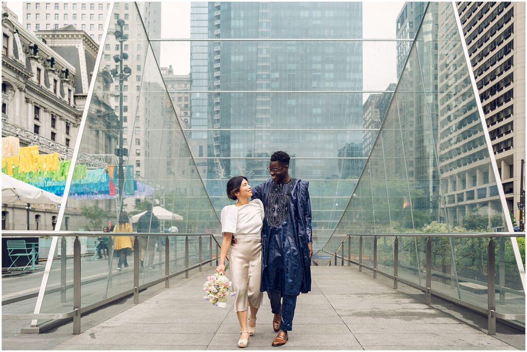 A bride and groom walk between glass walls near City Hall Philadelphia.