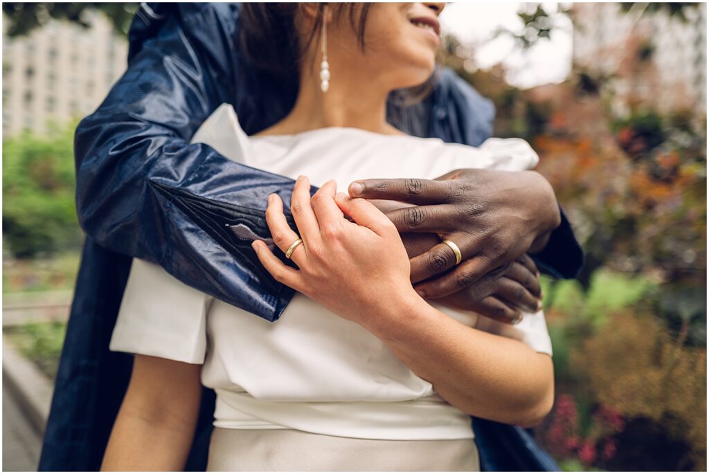 A groom puts his arms around a bride's shoulders, showing their wedding rings.