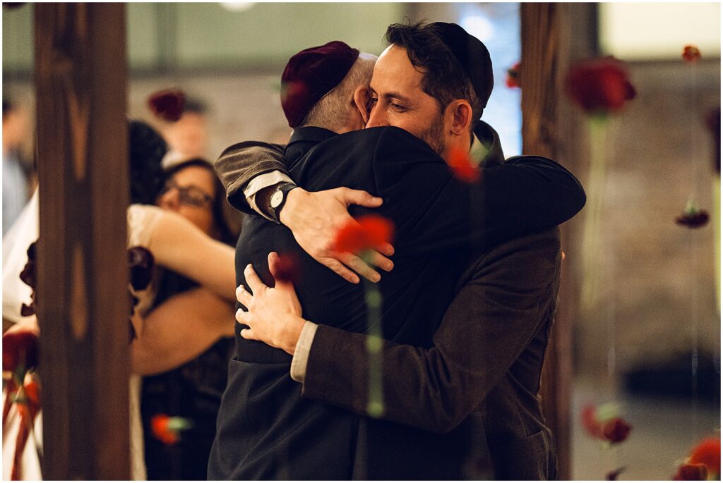 A groom hugs his father in law beside a floral chuppah.