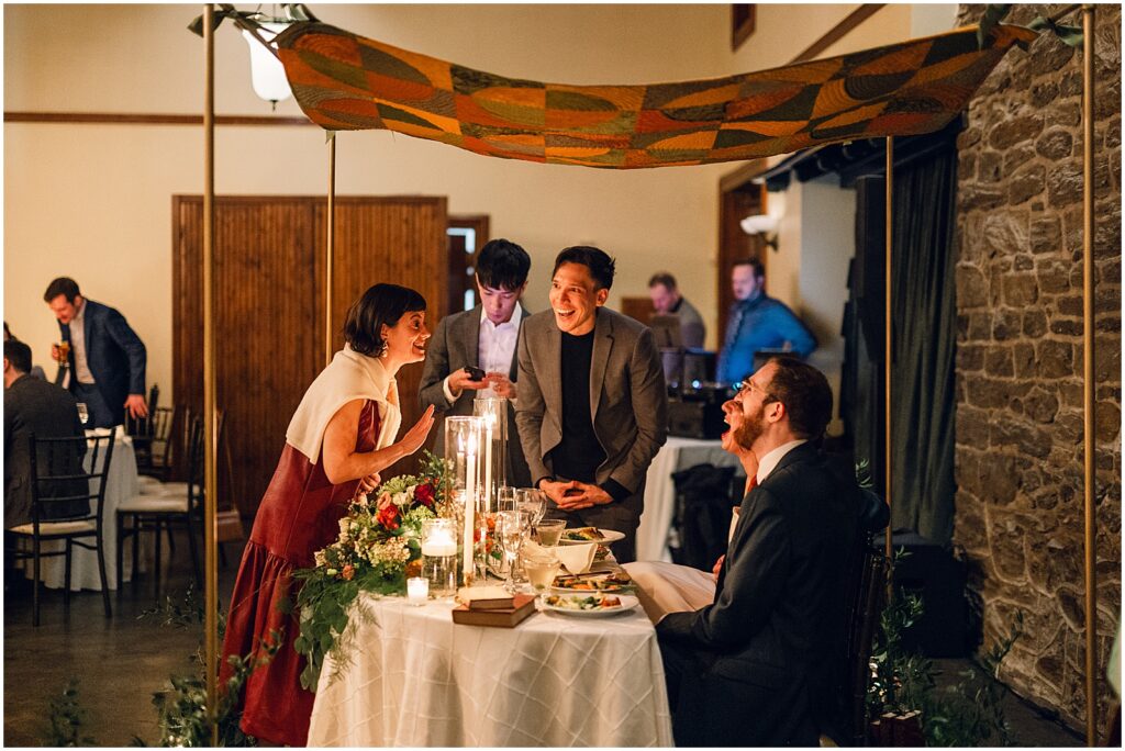 A guest leans over a candlelit sweetheart table to talk to a bride and groom.