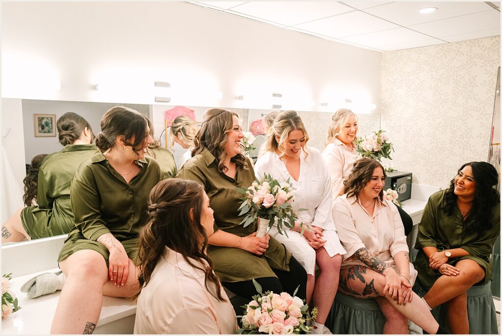 A bride and her bridesmaids sit in robes in front of a makeup table.