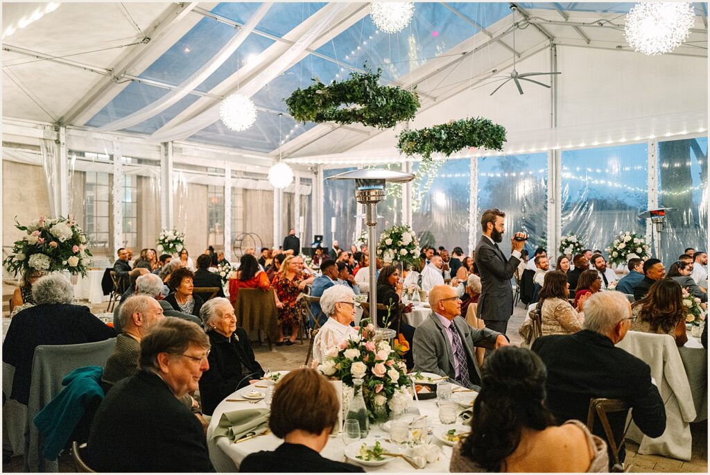 Wedding guests sit at tables and listen to a toast.