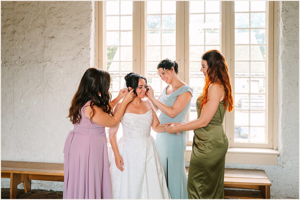 Bridesmaids in colorful dresses help a bride put on her jewelry.