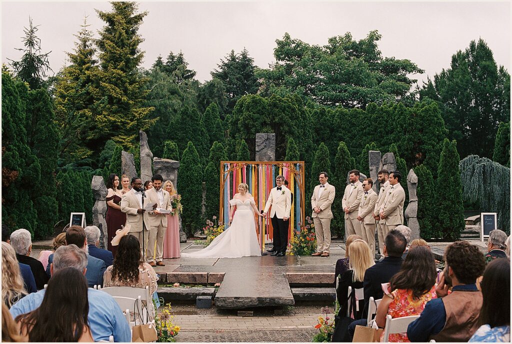 A bride and groom hold hands while a groomsmen does a reading.