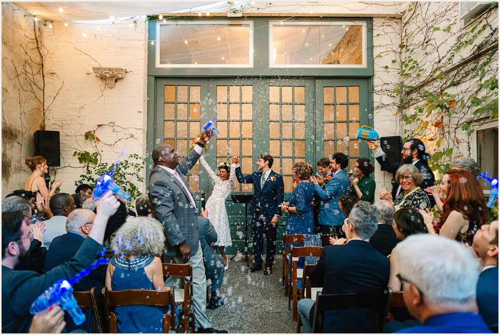A bride and groom cheer with wedding guests as the guests blow bubbles.