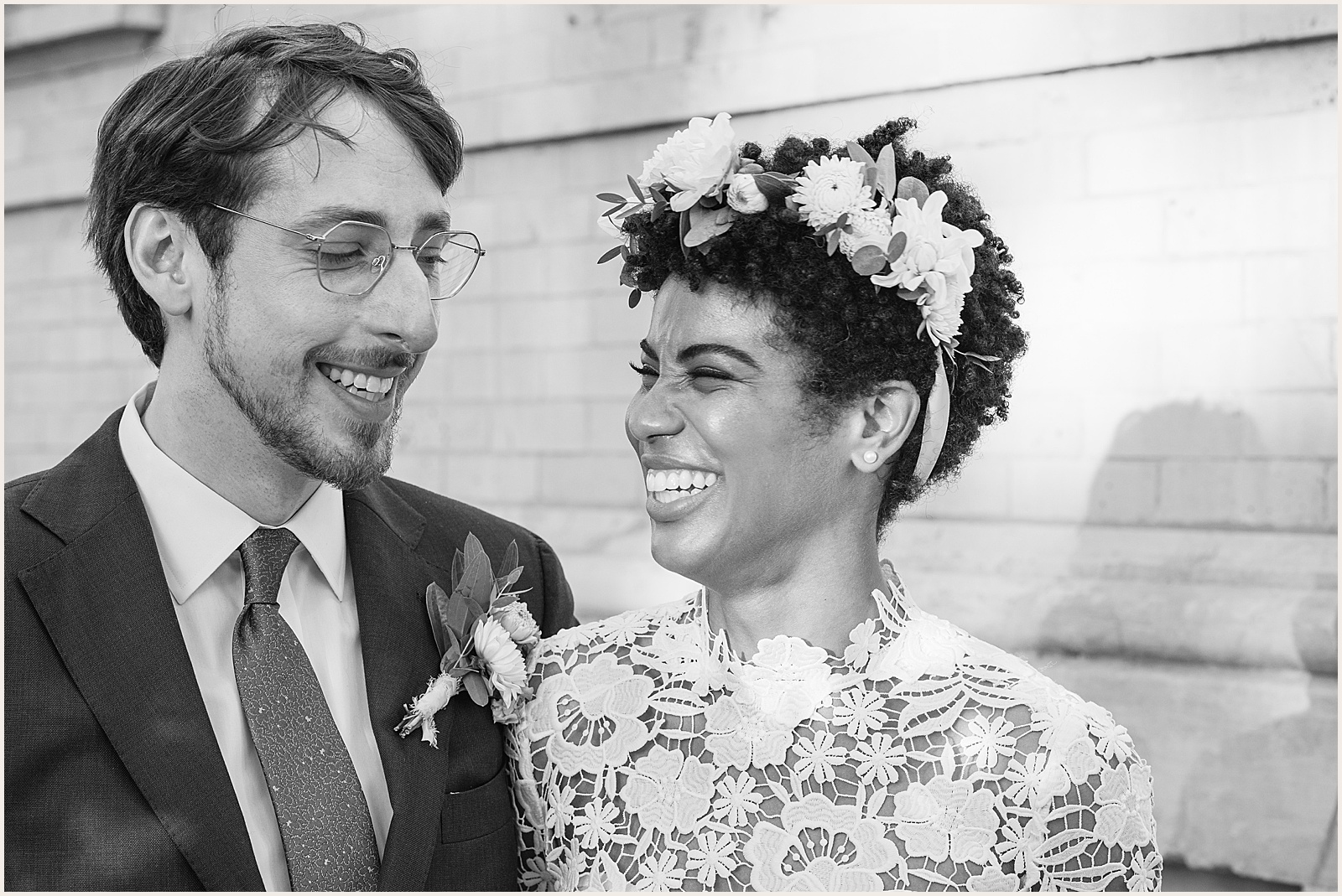 A bride and groom laugh in front of a white brick wall.