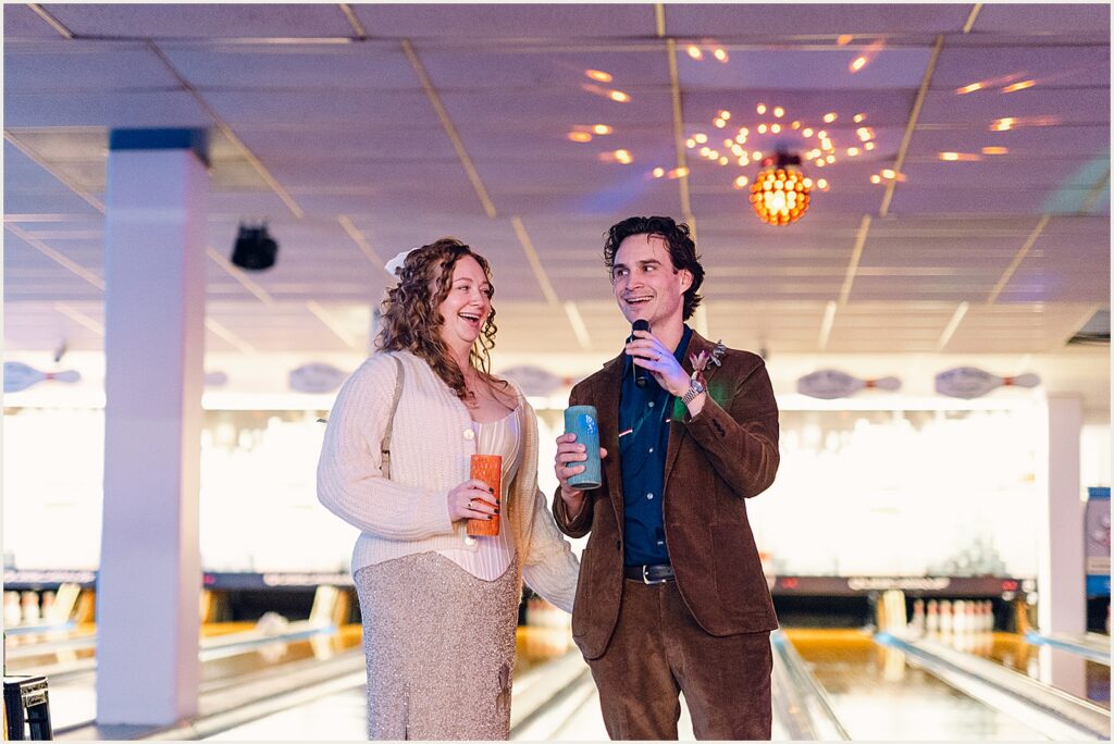 A bride and groom stand at a bowling lane laughing.