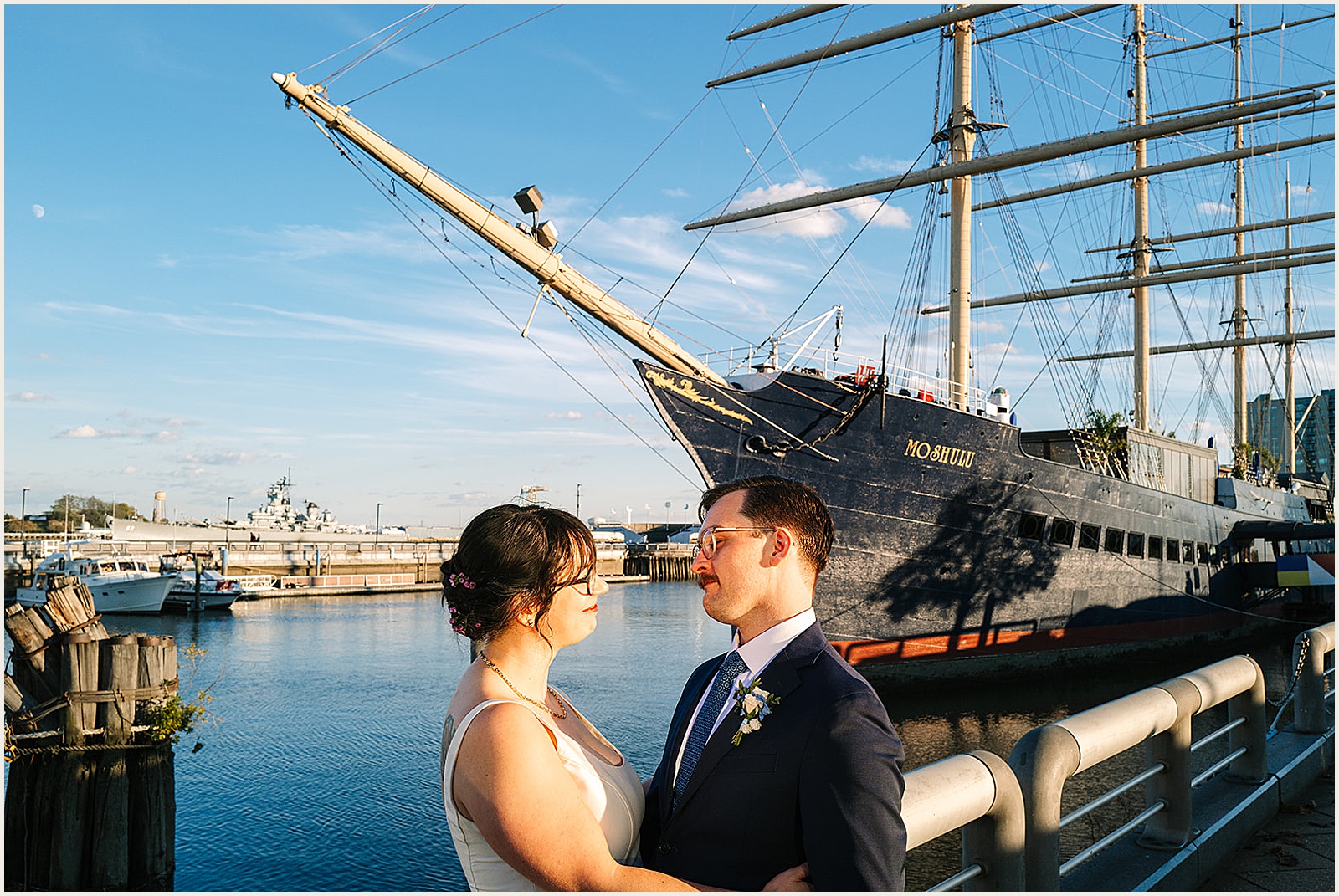 A bride and groom embrace on the sidewalk with Moshulu Philadelphia behind them.